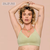 A woman wearing a light sage green nursing bra with the brand name 'leash.' visible on the band, posing with her arms raised against a solid pale pink background.