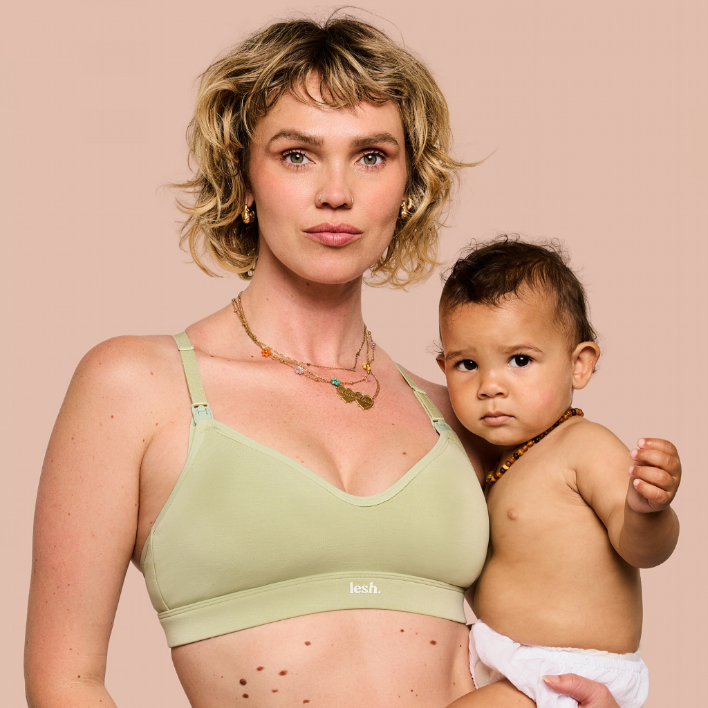 A woman holding a baby wearing a light sage green nursing bra with the brand name 'lesh.' visible on the band, against a solid pale pink background.