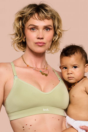 A woman holding a baby wearing a light sage green nursing bra with the brand name 'lesh.' visible on the band, against a solid pale pink background.