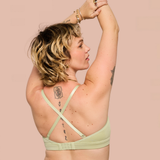 A woman with short blonde hair is shown from the back wearing a light sage green bra with crisscross straps, posing with her arms raised against a solid pale pink background.