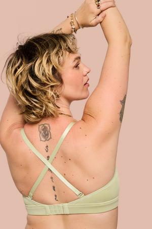 A woman with short blonde hair is shown from the back wearing a light sage green bra with crisscross straps, posing with her arms raised against a solid pale pink background.