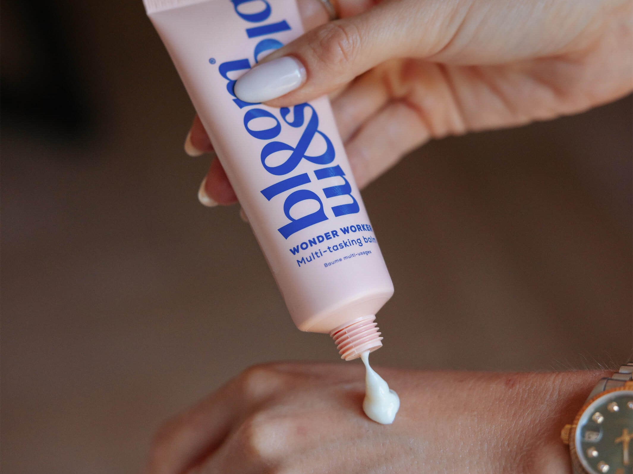 A close-up of a person squeezing a pink tube of lip & cheek tint onto the back of their hand. The tube has blue text that reads 'lip & cheek' and 'WONDER WORLD MULTI-REVIEW TINT'.