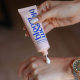 A close-up of a person squeezing a pink tube of lip & cheek tint onto the back of their hand. The tube has blue text that reads 'lip & cheek' and 'WONDER WORLD MULTI-REVIEW TINT'.