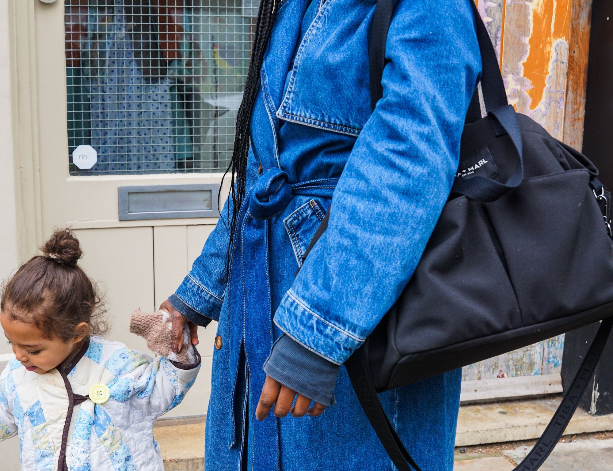 Woman in a blue denim coat holding a black bag, with a child next to her, outdoors.