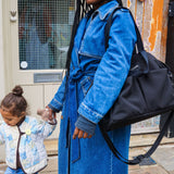 Woman in a blue denim coat holding a black bag, with a child next to her, outdoors.