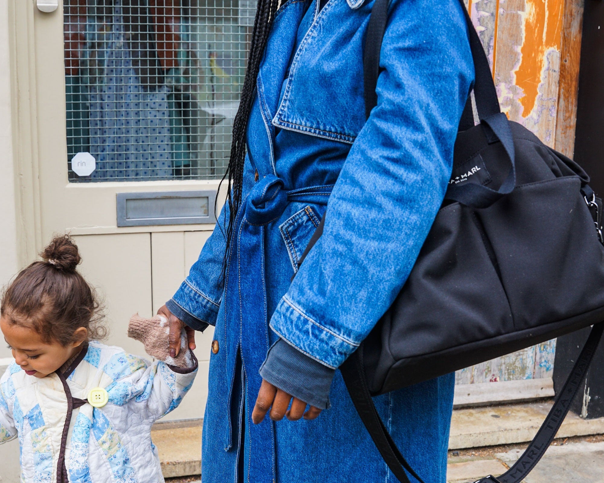 Woman in a blue denim coat holding a black bag, with a child next to her, outdoors.