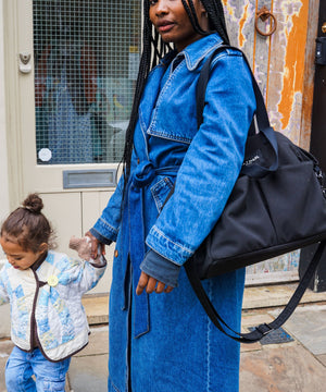Woman in a blue denim coat holding a black bag, with a child next to her, outdoors.