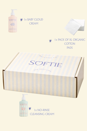 A Softie gift set on a pale cream background, featuring a striped cardboard box labeled SOFTIE, a pump bottle of Baby Cloud Cream, a pack of organic cotton pads, and a bottle of No-Rinse Cleansing Cream with text callouts around it.