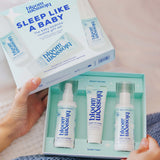 A person holds an open mint-green Blossom Sleep Like a Baby routine gift set box, showing three baby bedding products inside: a spray bottle, a tube, and a pump bottle, with the outer box lid displayed above.