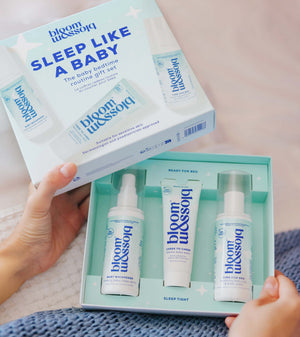 A person holds an open mint-green Blossom Sleep Like a Baby routine gift set box, showing three baby bedding products inside: a spray bottle, a tube, and a pump bottle, with the outer box lid displayed above.