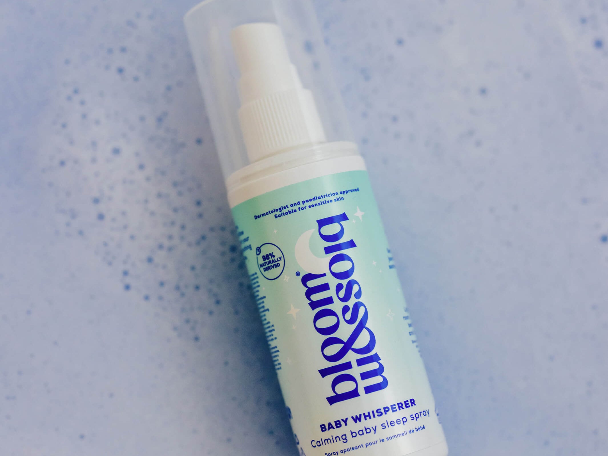 A bottle of bloom babywhisperer baby shower spray with a white pump and a light blue-green label, shown on a textured light blue background.