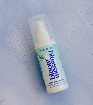 A bottle of bloom babywhisperer baby shower spray with a white pump and a light blue-green label, shown on a textured light blue background.