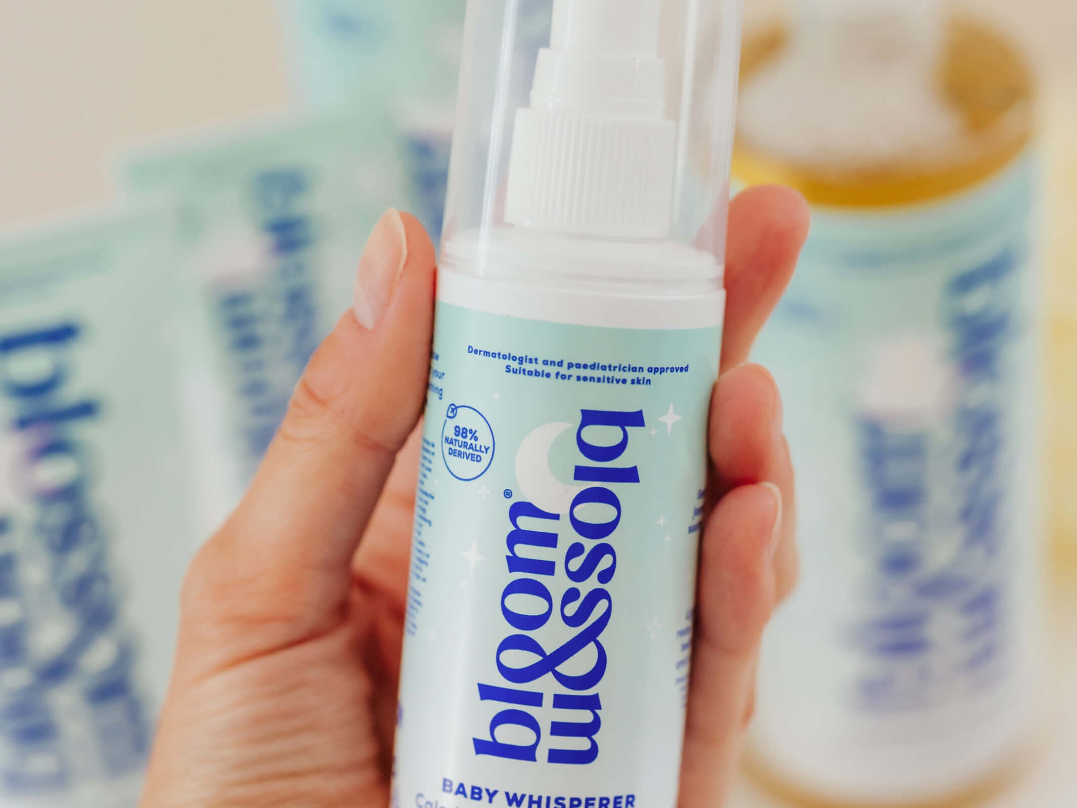 A hand holding a bottle of bloom & assolo Baby Whisperer Coloring Baby Sleep Spray, with other bottles blurred in the background.