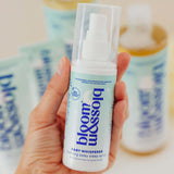A hand holding a bottle of bloom & assolo Baby Whisperer Coloring Baby Sleep Spray, with other bottles blurred in the background.