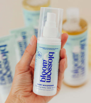 A hand holding a bottle of bloom & assolo Baby Whisperer Coloring Baby Sleep Spray, with other bottles blurred in the background.