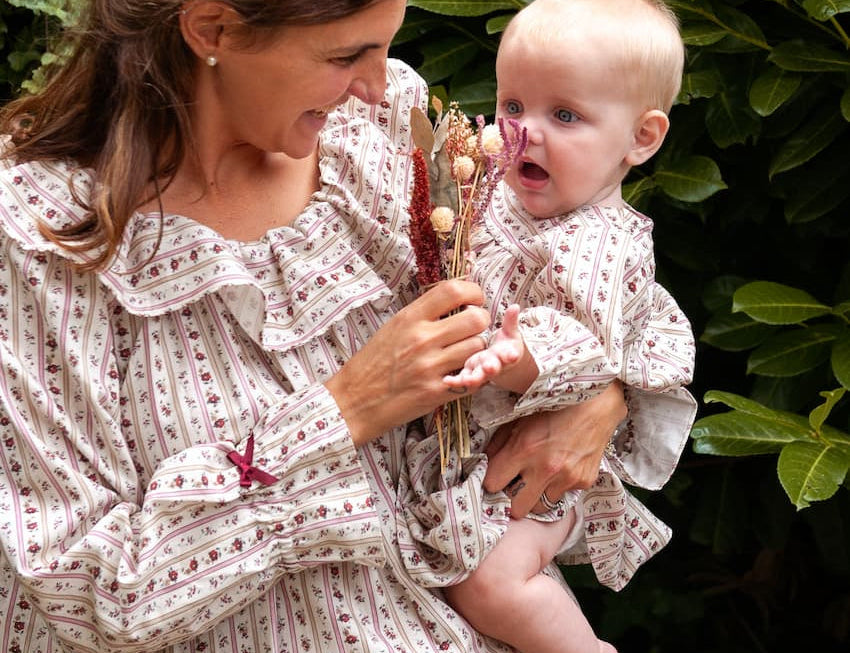 Beatrice Rose Stripe nightie worn by a woman holding a baby in a matching outfit and dried flowers by a hedge