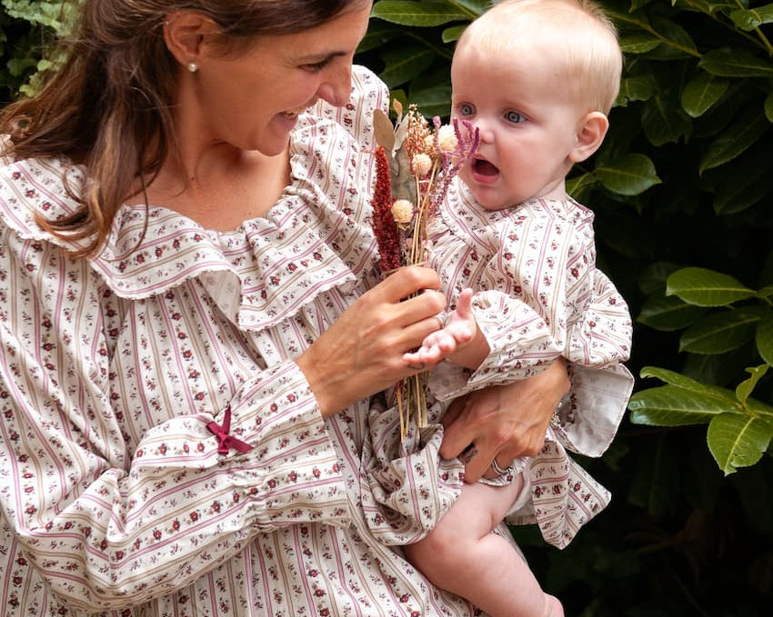 Beatrice Rose Stripe nightie worn by a woman holding a baby in a matching outfit and dried flowers by a hedge