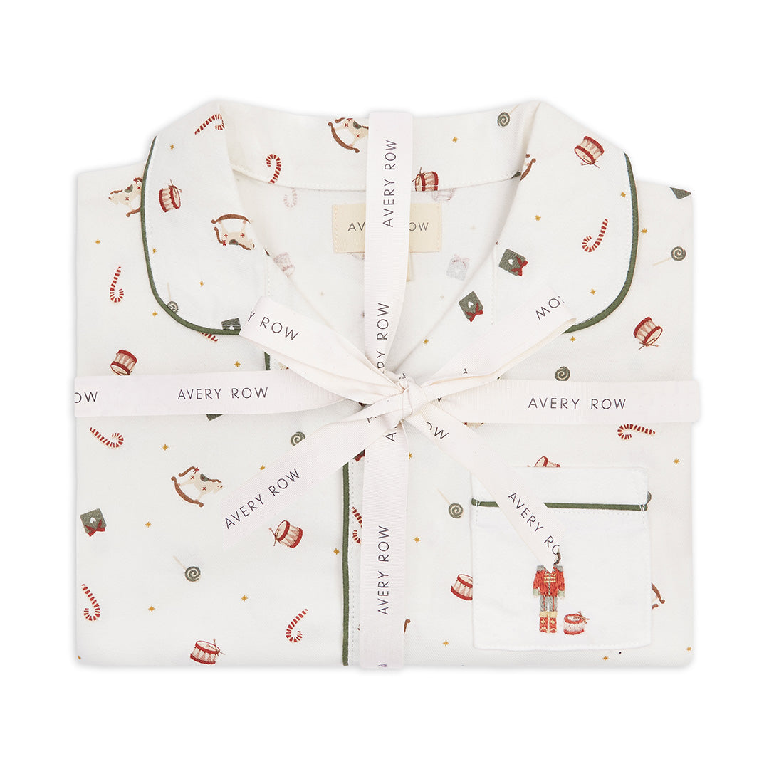 A folded white pajama set with a festive holiday print, tied with a white ribbon printed with the brand name 'Avery Row'. The pajamas feature a pattern of red and gold ornaments, candy canes, and a small nutcracker on the pocket, with dark green piping.