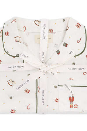 A folded white pajama set with a festive holiday print, tied with a white ribbon printed with the brand name 'Avery Row'. The pajamas feature a pattern of red and gold ornaments, candy canes, and a small nutcracker on the pocket, with dark green piping.