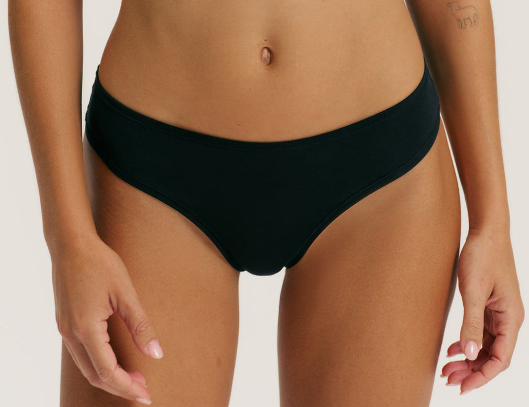 A close-up of a model wearing a solid black bikini bottom against a clean, light off-white background.