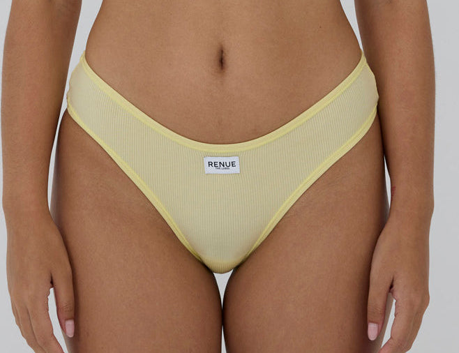 A close-up of a model wearing a pale yellow ribbed bikini bottom with a white brand label reading 'RENUE' on the front, set against a plain light gray background.