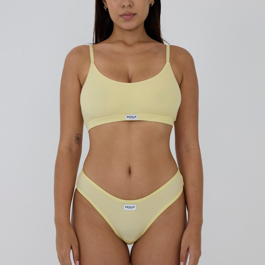 A model wearing a pale yellow two-piece bikini set with a scoop neck top and matching high-cut bottoms, featuring a small white brand label that reads 'MENSTRU'.