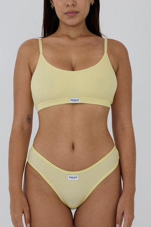 A model wearing a pale yellow two-piece bikini set with a scoop neck top and matching high-cut bottoms, featuring a small white brand label that reads 'MENSTRU'.
