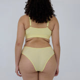 A woman shown from the back wearing a pale yellow two-piece bikini set against a plain light gray studio background.