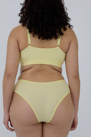 A woman shown from the back wearing a pale yellow two-piece bikini set against a plain light gray studio background.