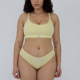A model wearing a pale yellow two-piece bikini set with a scoop neck top and matching high-cut bottoms, featuring a small white brand label that reads 'MENDIA'.