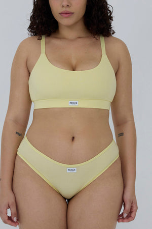 A model wearing a pale yellow two-piece bikini set with a scoop neck top and matching high-cut bottoms, featuring a small white brand label that reads 'MENDIA'.