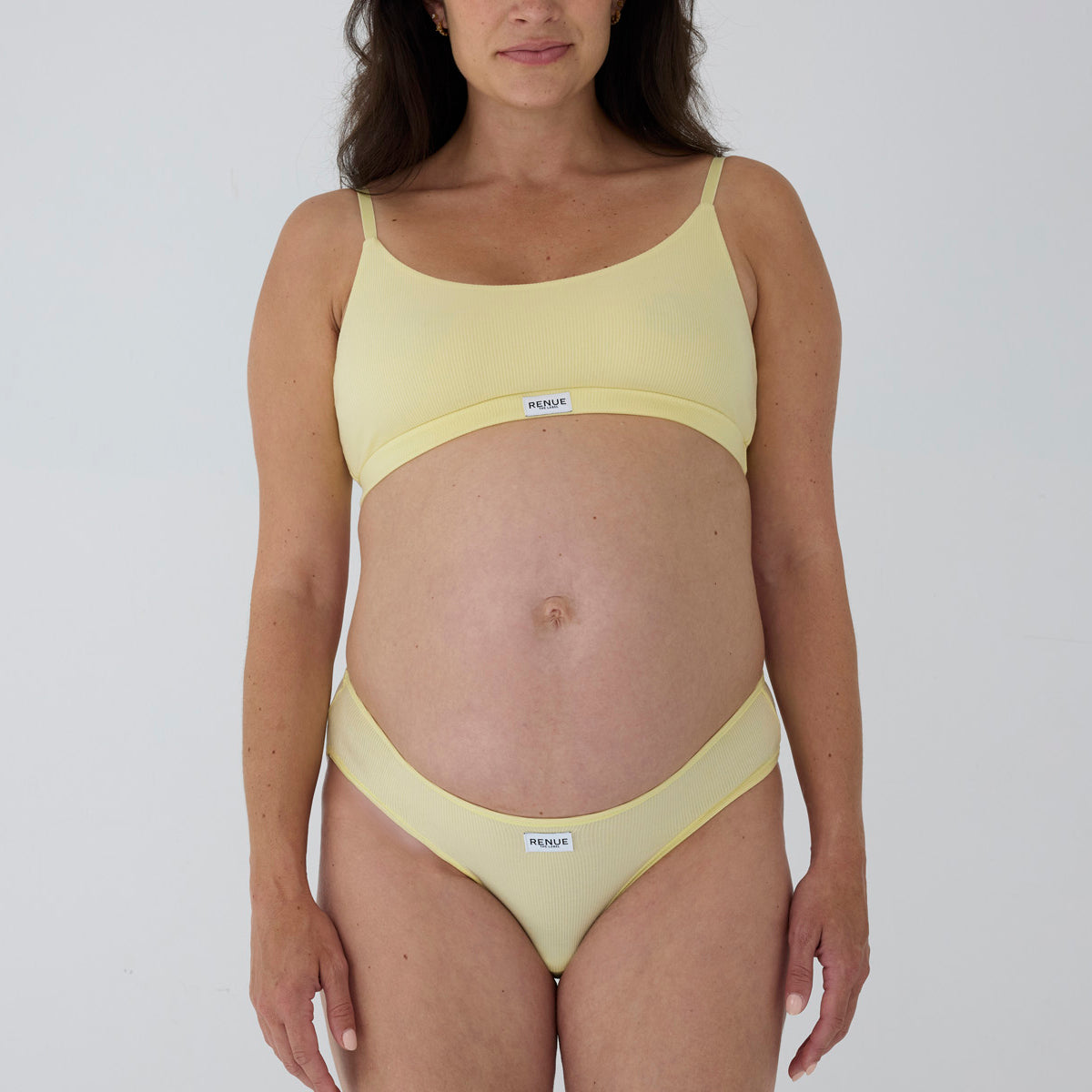 A pregnant model wearing a pale yellow two-piece bikini set with a scoop neck top and matching high-cut bottoms, featuring a small white brand label that reads 'MAGNA'.
