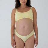 A pregnant model wearing a pale yellow two-piece bikini set with a scoop neck top and matching high-cut bottoms, featuring a small white brand label that reads 'MAGNA'.