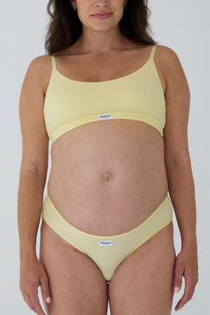 A pregnant model wearing a pale yellow two-piece bikini set with a scoop neck top and matching high-cut bottoms, featuring a small white brand label that reads 'MAGNA'.