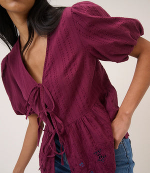 Woman wearing a burgundy open-knit cardigan over a black top with blue jeans on a plain background