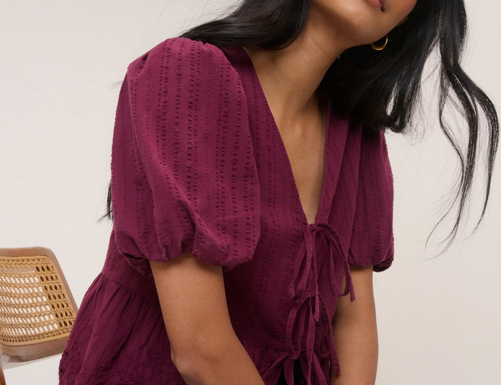 Woman wearing a burgundy top with puffed sleeves and blue jeans sitting against a white wall.