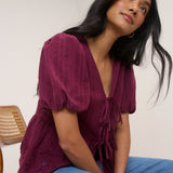 Woman wearing a burgundy top with puffed sleeves and blue jeans sitting against a white wall.
