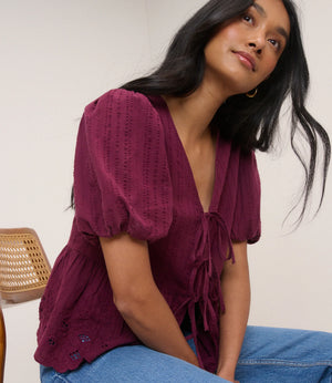 Woman wearing a burgundy top with puffed sleeves and blue jeans sitting against a white wall.