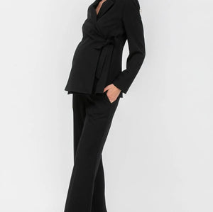 A woman modeling a black long-sleeve wrap-style blazer with a tie waist and matching black wide-leg trousers, paired with taupe high heels, against a plain light gray studio background.