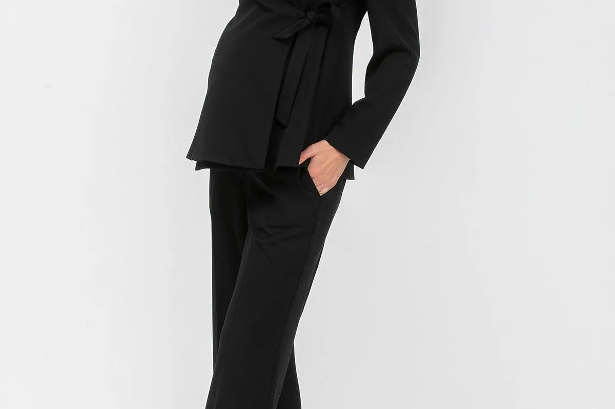 A woman modeling a black long-sleeve wrap-style blazer with a tie waist and matching black wide-leg trousers, paired with taupe high heels, against a plain light gray studio background.