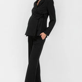 A woman modeling a black long-sleeve wrap-style blazer with a tie waist and matching black wide-leg trousers, paired with taupe high heels, against a plain light gray studio background.