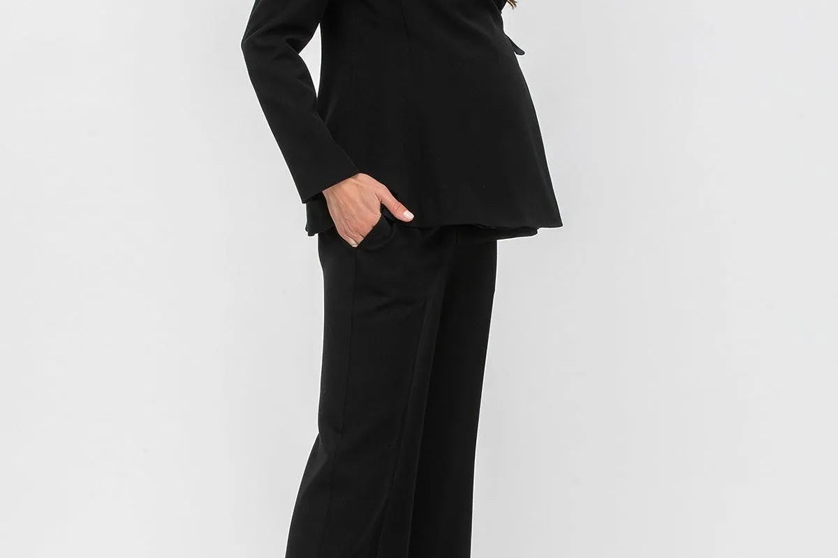 A woman modeling a black two-piece suit consisting of a structured blazer with notched lapels and matching wide-leg trousers, paired with taupe pointed-toe heels against a light gray studio background.