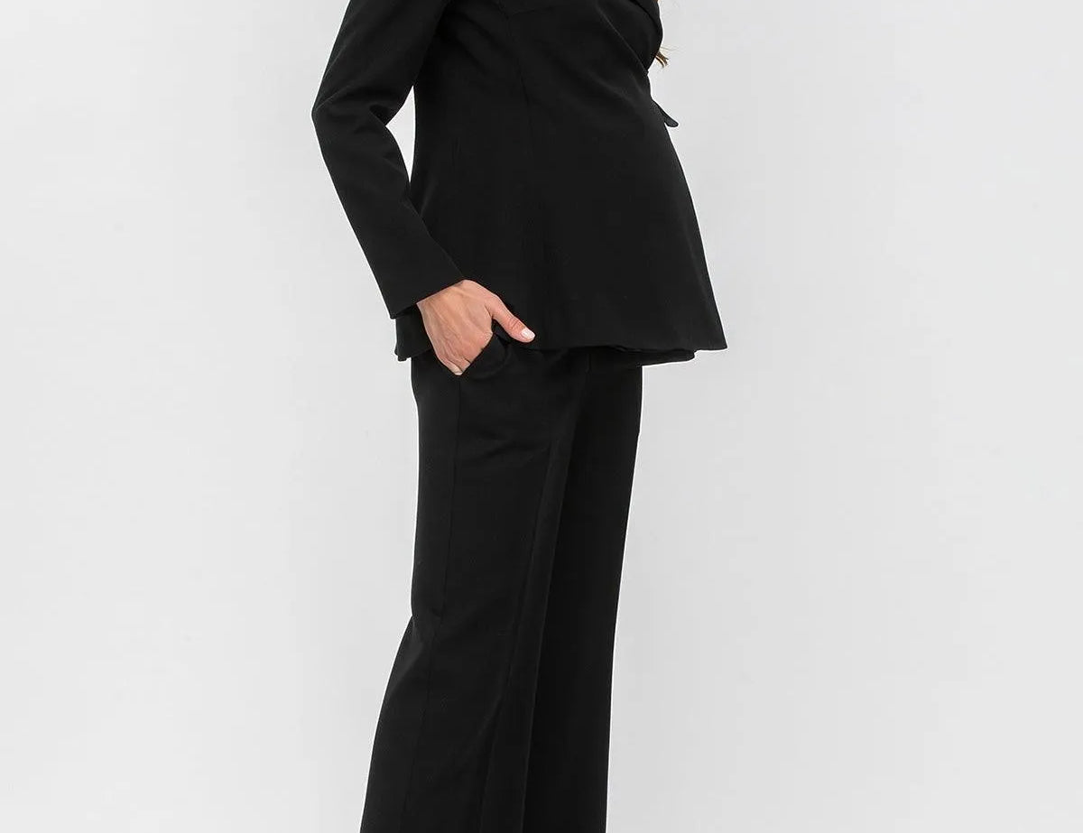 A woman modeling a black two-piece suit consisting of a structured blazer with notched lapels and matching wide-leg trousers, paired with taupe pointed-toe heels against a light gray studio background.