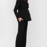 A woman modeling a black two-piece suit consisting of a structured blazer with notched lapels and matching wide-leg trousers, paired with taupe pointed-toe heels against a light gray studio background.