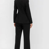 A woman modeling a black tailored pantsuit from the back, featuring a structured blazer with a high collar and matching wide-leg trousers, set against a plain light gray studio background.