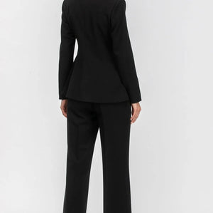 A woman modeling a black tailored pantsuit from the back, featuring a structured blazer with a high collar and matching wide-leg trousers, set against a plain light gray studio background.