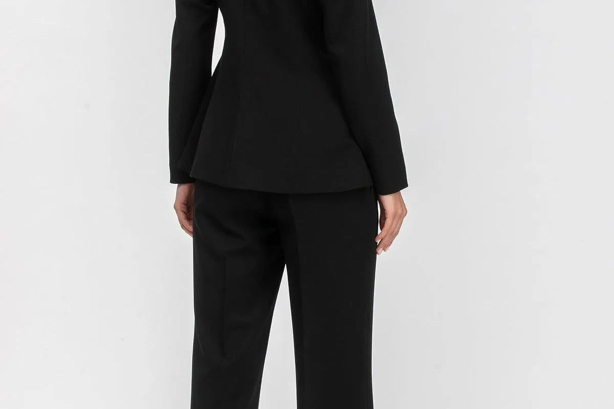 A woman modeling a black tailored pantsuit from the back, featuring a structured blazer with a high collar and matching wide-leg trousers, set against a plain light gray studio background.