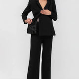 A woman modeling a black tailored pantsuit consisting of a structured blazer with a wrap-style front and matching wide-leg trousers, paired with pointed heels against a light gray studio background.