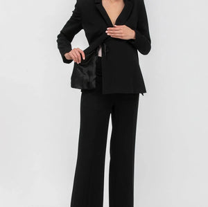 A woman modeling a black tailored pantsuit consisting of a structured blazer with a wrap-style front and matching wide-leg trousers, paired with pointed heels against a light gray studio background.