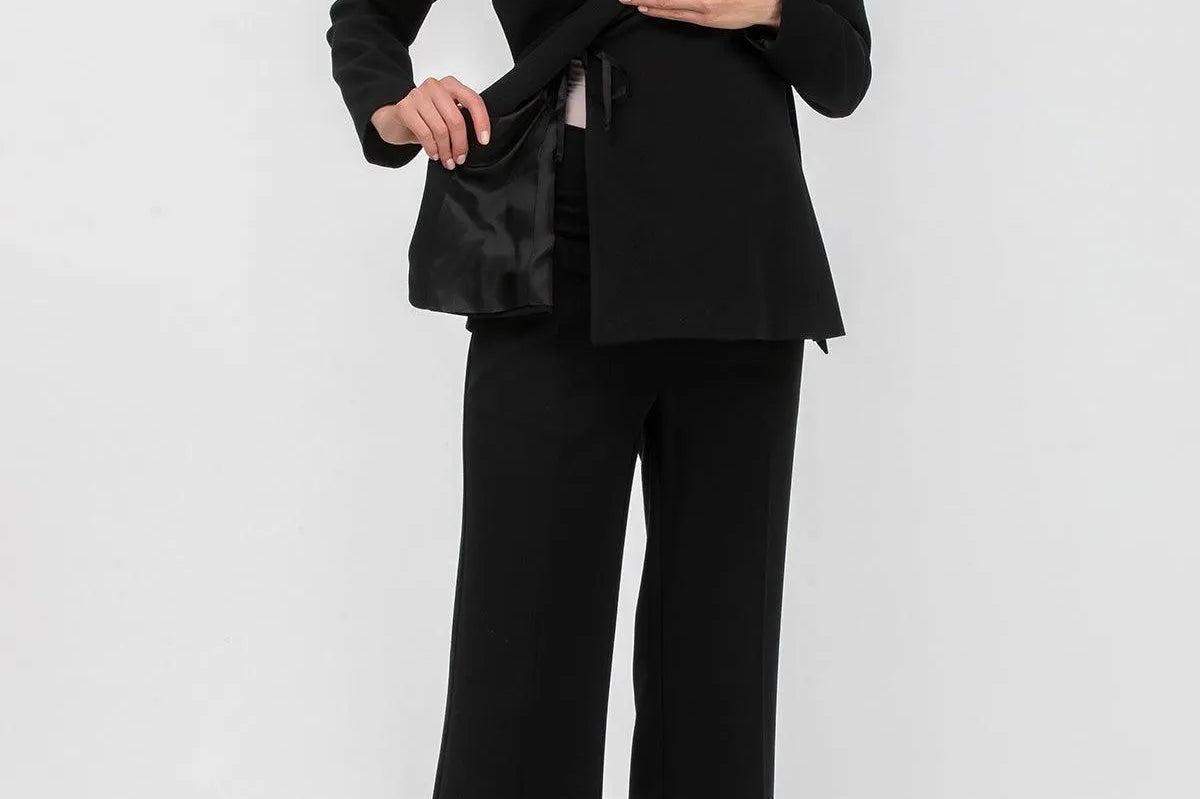 A woman modeling a black tailored pantsuit consisting of a structured blazer with a wrap-style front and matching wide-leg trousers, paired with pointed heels against a light gray studio background.